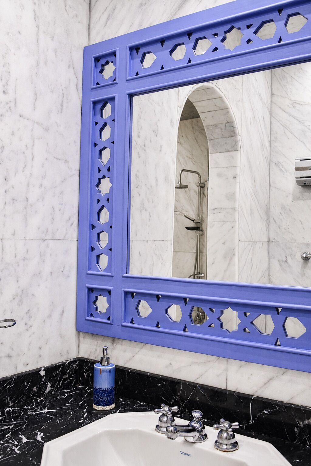 A bathroom with a marble wall, a blue-framed mirror, a sink with two faucets, and a shower area.