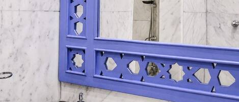 A bathroom with a marble wall, a blue-framed mirror, a sink with two faucets, and a shower area.
