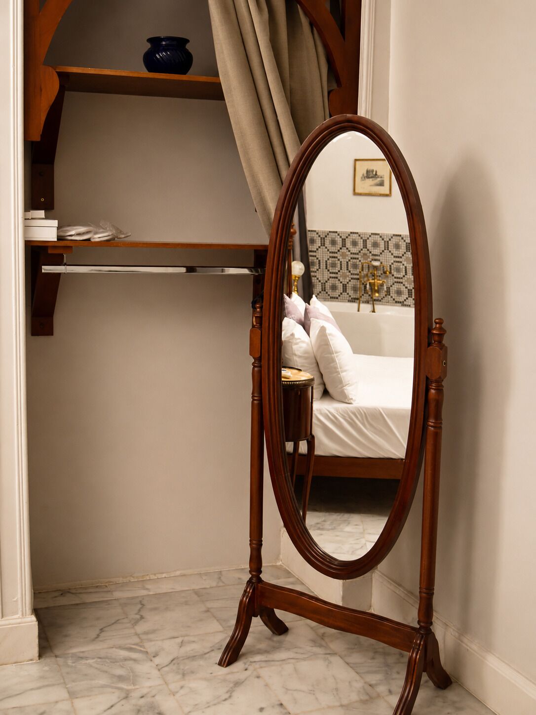 A corner room with a wooden floor, a bed with white bedding, a wooden dresser, a mirror, and a shelf with a bowl and books.