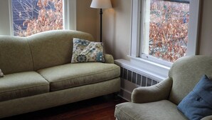 Een woonkamer met een bank en fauteuil, een vloerlamp en een raam waar herfstbladeren te zien zijn.