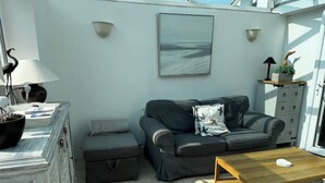 Un espacio habitable pequeño con un sofá gris, un otoman pequeño, una mesa de centro de madera y un estante en la pared con objetos decorativos.