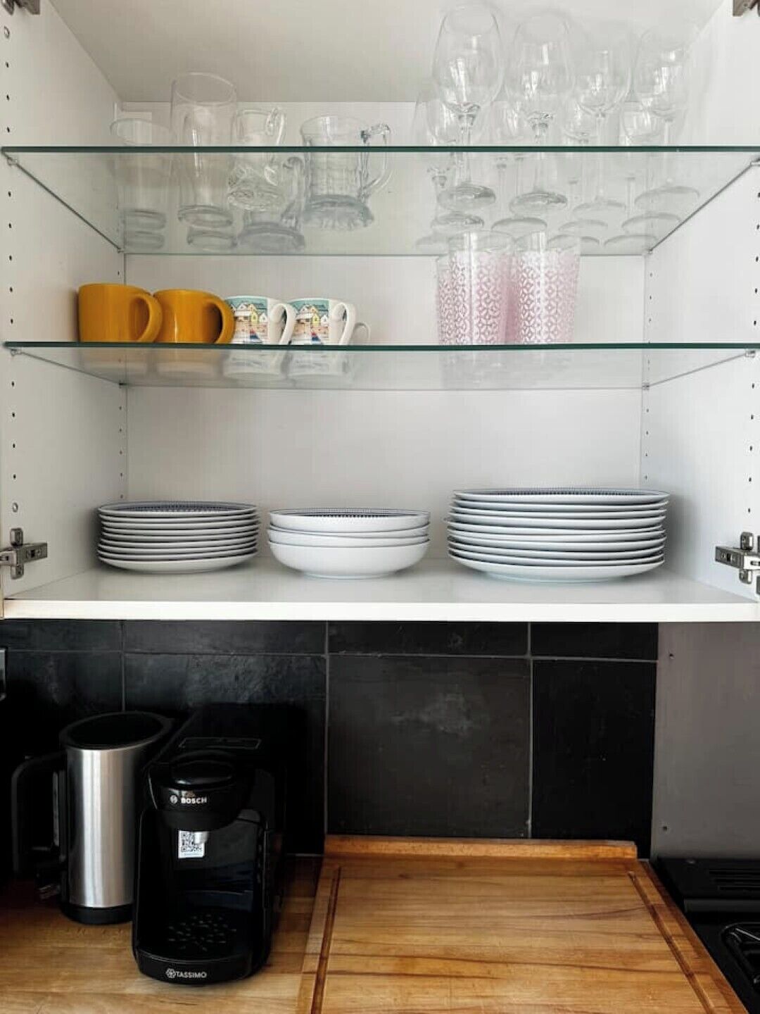 A kitchen with a glass shelf holding various glassware, a coffee maker, and stacked plates.