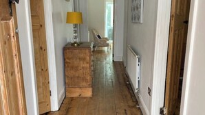Un pasillo con piso de madera, paredes blancas y un candelabro. Se observa un aparador de madera con una lámpara, y una silla al fondo.