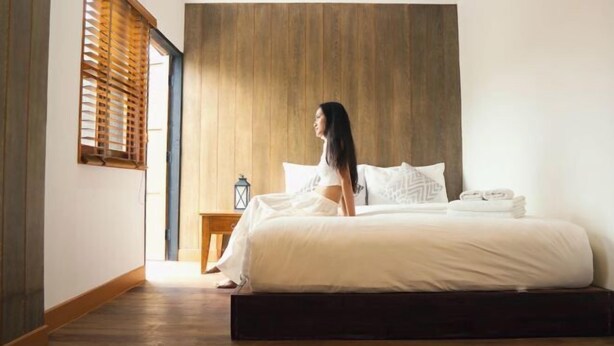Un dormitorio con cabecera de madera, cama con ropa blanca, mesita de noche con una linterna y ventana con persianas de madera.
