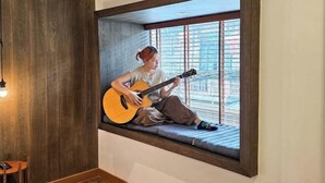 Una persona toca la guitarra en un asiento junto a la ventana, con vistas a edificios.