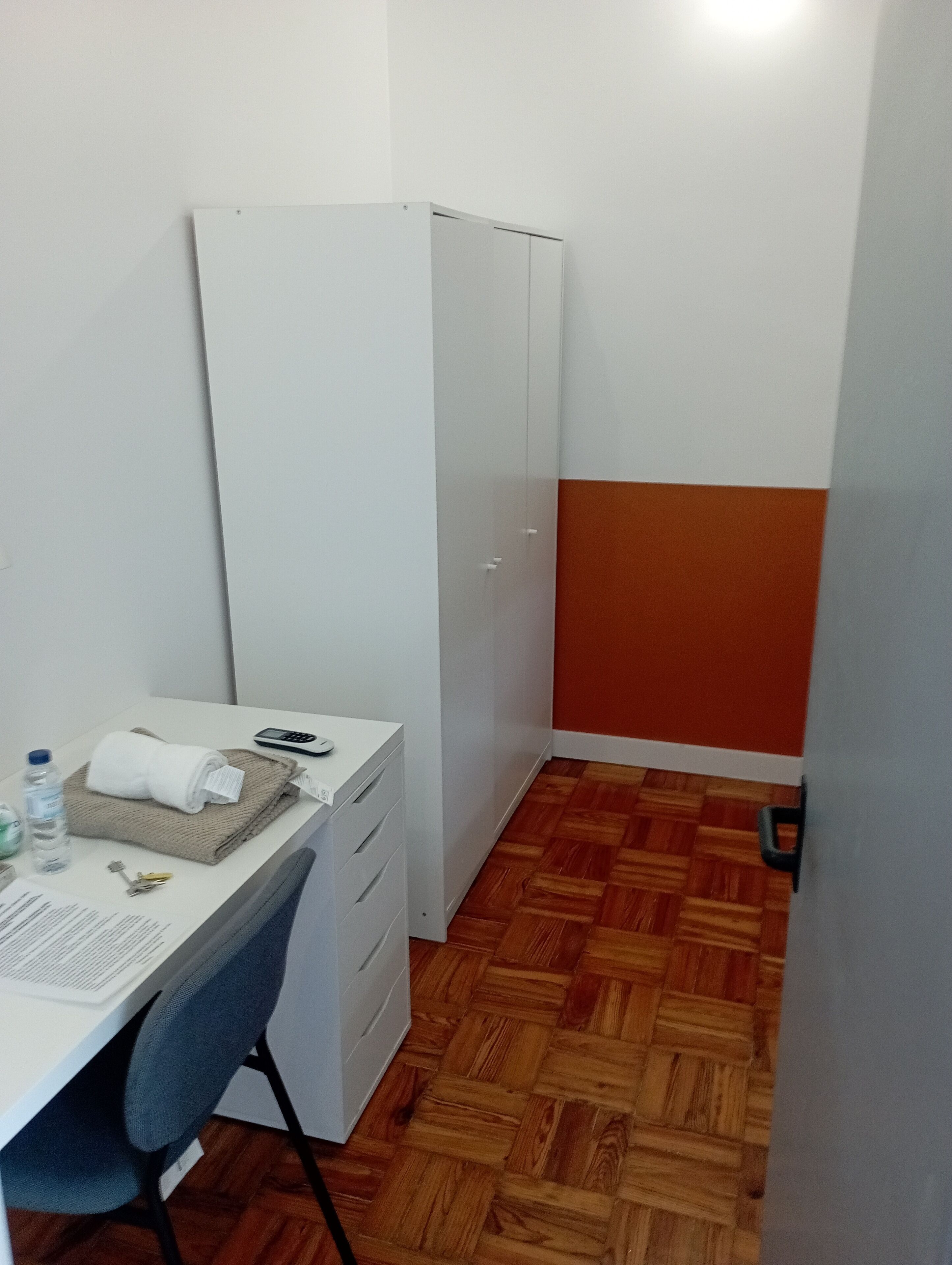A small room with a white desk, a chair, a wardrobe, and a wooden parquet floor.