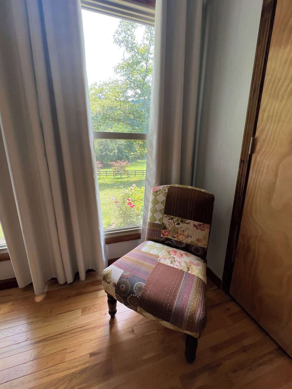 Ein Stuhl mit einem floralen Kissen in einem Raum mit Holzboden und Blick auf einen Garten durch ein Fenster mit weißen Vorhängen.