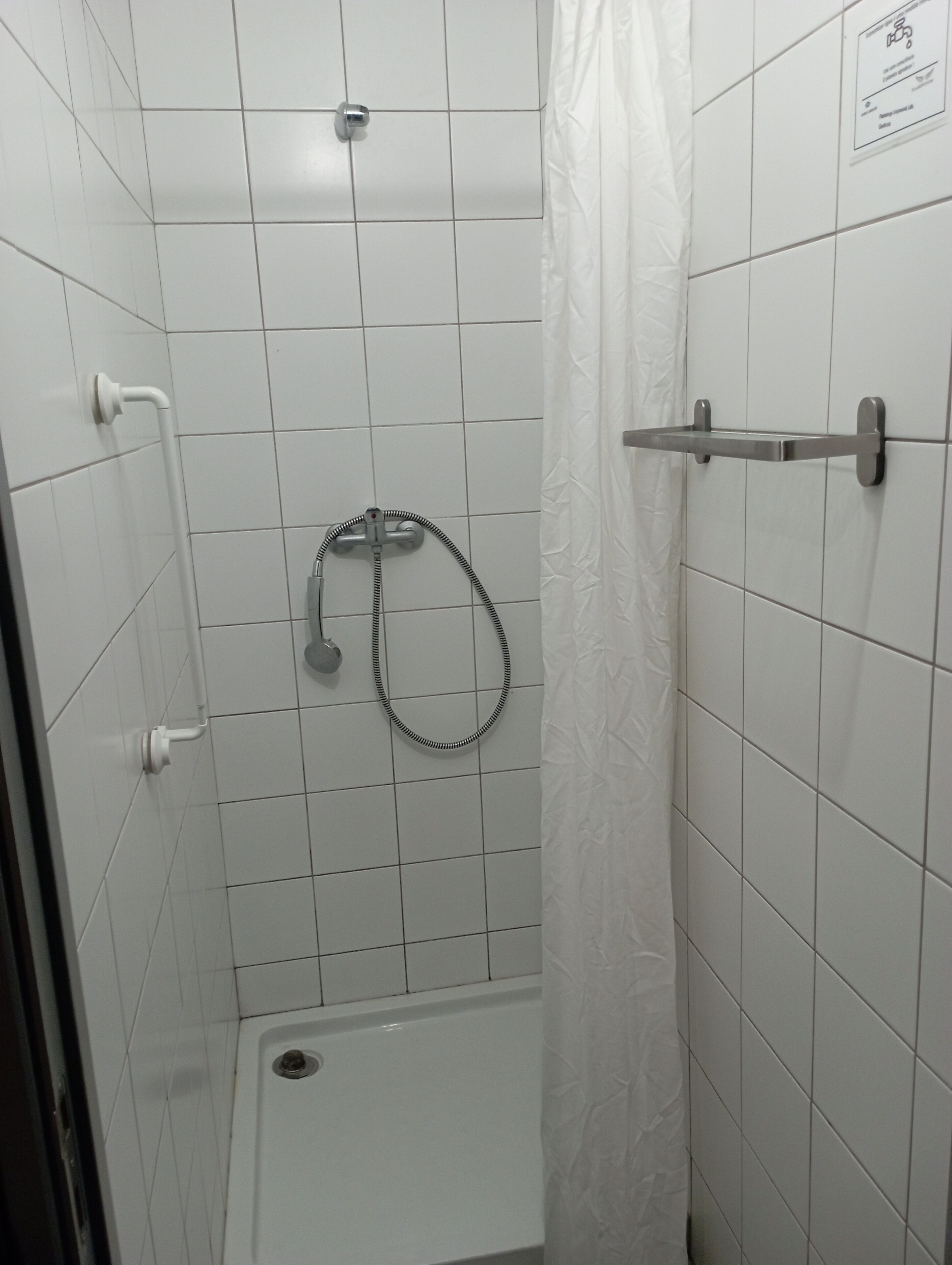 Um chuveiro pequeno revestido de azulejos brancos, com um chuveiro manual, um porta-toalhas e uma porta com puxador.