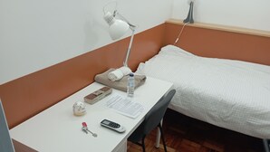 Een kleine, netjes ingerichte slaapkamer met een wit bureau, een bed met een witte dekbedovertrek, een lamp, een fles water, zakdoekjes, een sleutel en een mobiele telefoon.