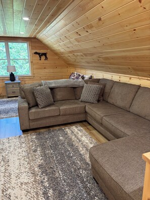 A cozy room with a sectional sofa, a small table, and a window with a view of greenery. - New Built! 2025 Cozy 3-bedroom chalet with water access to Rangeley Lake (Rangeley)