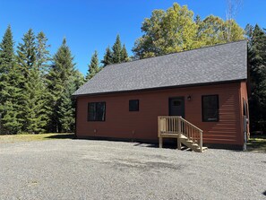 A single-story building with a gravel driveway, a wooden porch, and a metal door. - New Built! 2025 Cozy 3-bedroom chalet with water access to Rangeley Lake (Rangeley)