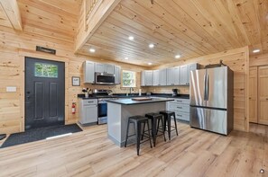 A modern kitchen with wooden walls, a central island, bar stools, stainless steel appliances, and a black door. - New Built! 2025 Cozy 3-bedroom chalet with water access to Rangeley Lake (Rangeley)