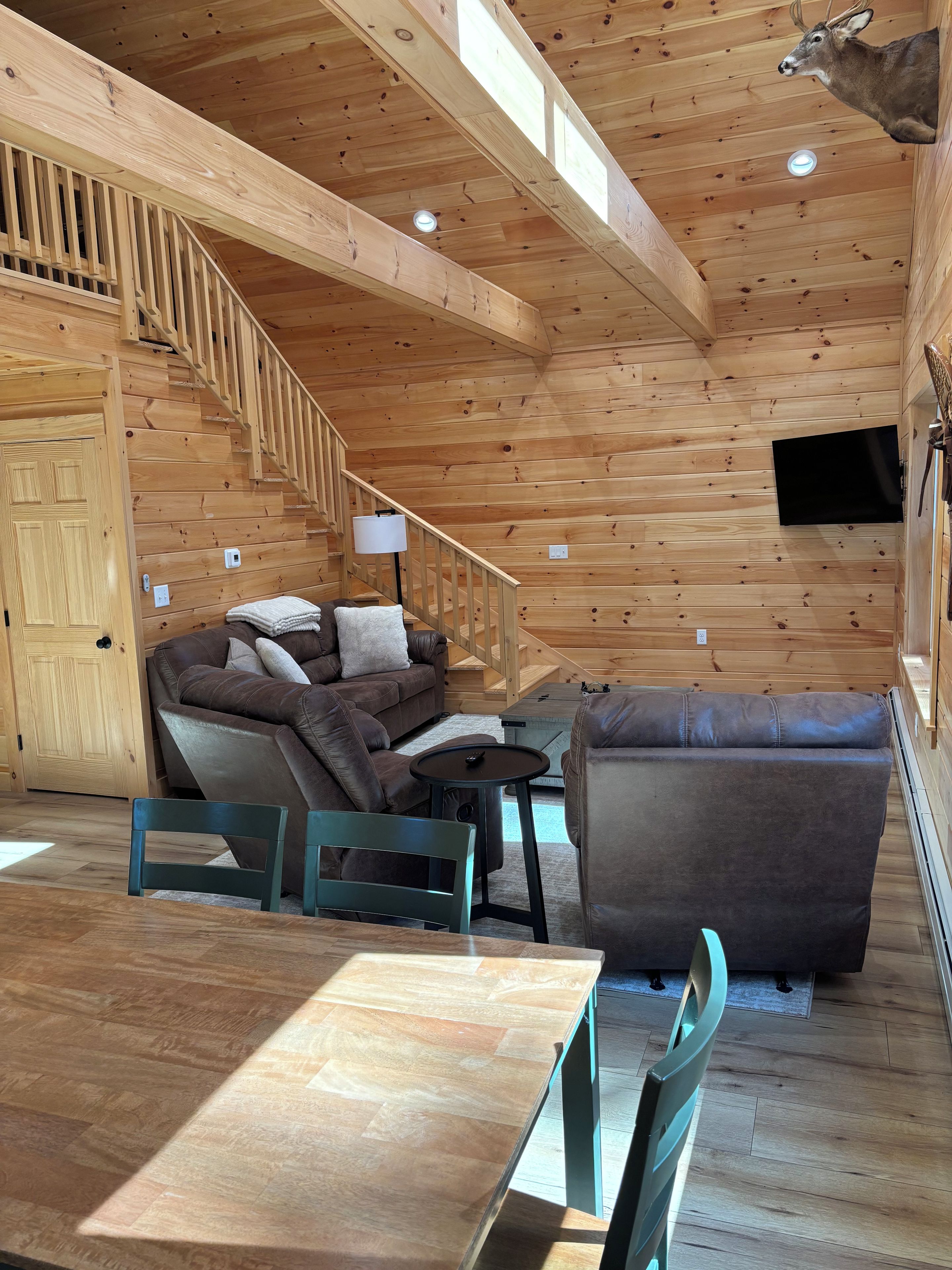 A wooden cabin interior with a sofa, a table, chairs, a staircase, and a mounted television.