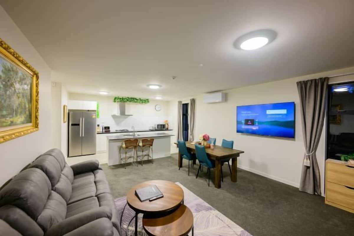 A living room with a sofa, a coffee table, a dining area with chairs, a TV, and a kitchen in the background.