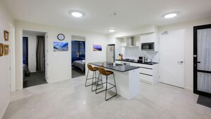 A modern kitchen with a central island, bar stools, and a microwave. There is a clock on the wall and a door leading to another room. - Stargazing Tekapo Apartment (Lake Tekapo)
