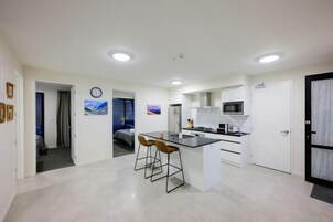 Una cocina moderna con una isla central, taburetes de bar y un microondas. Hay un reloj en la pared y una puerta que da a otra habitación.