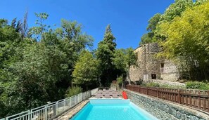 A swimming pool with a stone wall and a wooden fence, surrounded by trees and a stone building.