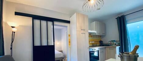 Habitación de hotel moderna con cama, zona de comedor, kitchenette y un amplio ventanal con cortinas.