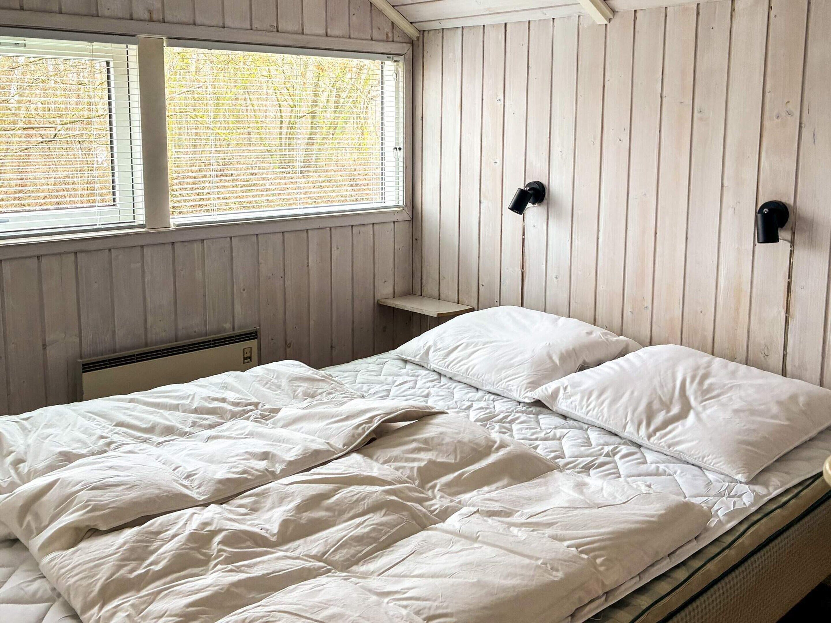 Makuuhuone, jossa on sänky, puuseinät, ikkuna, jossa on sälekaihtimet, ja seinään kiinnitetty valaisin.