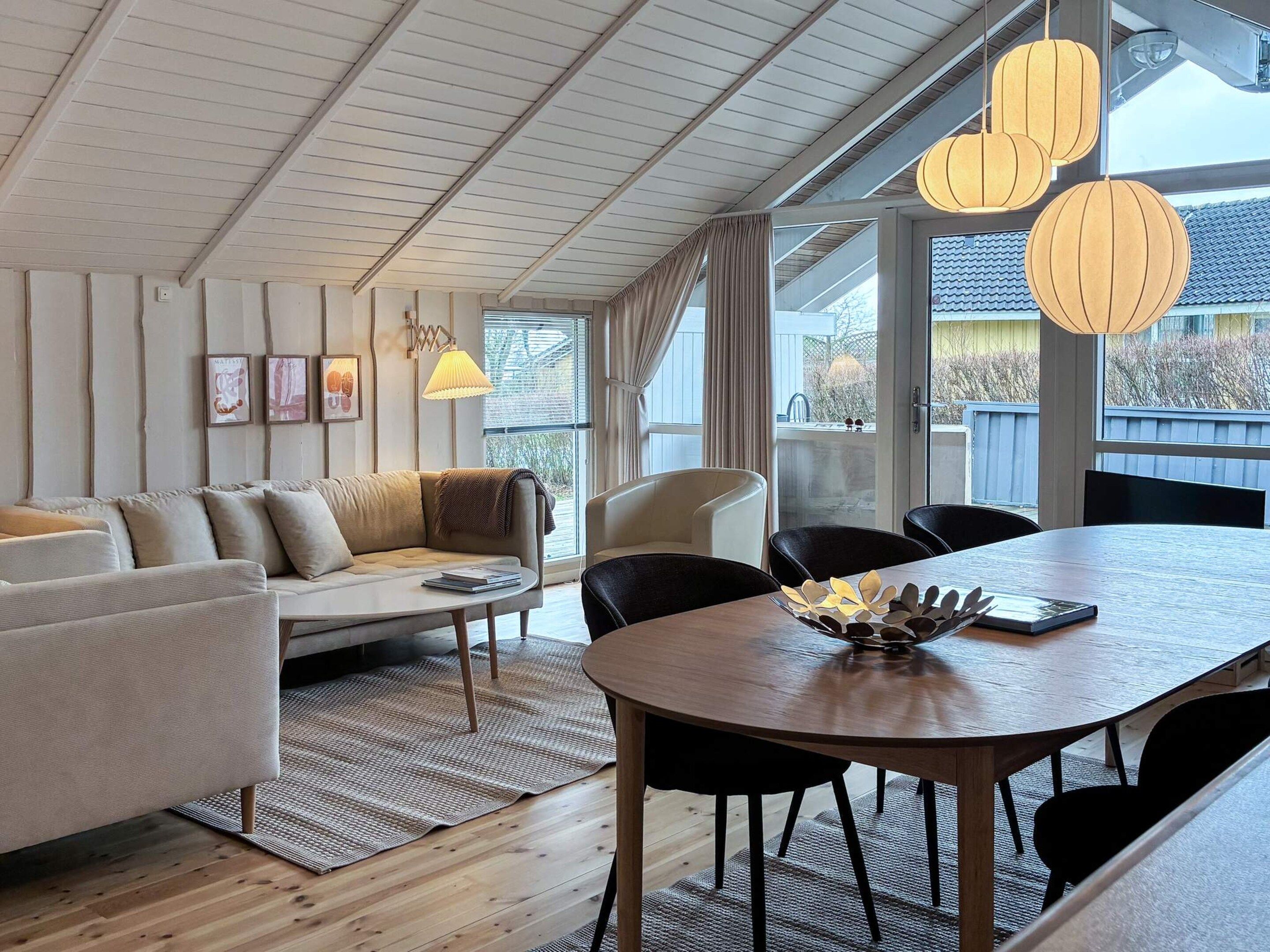 Una sala de estar moderna con sofá, mesa de comedor, sillas, ventanas amplias e iluminación colgante.