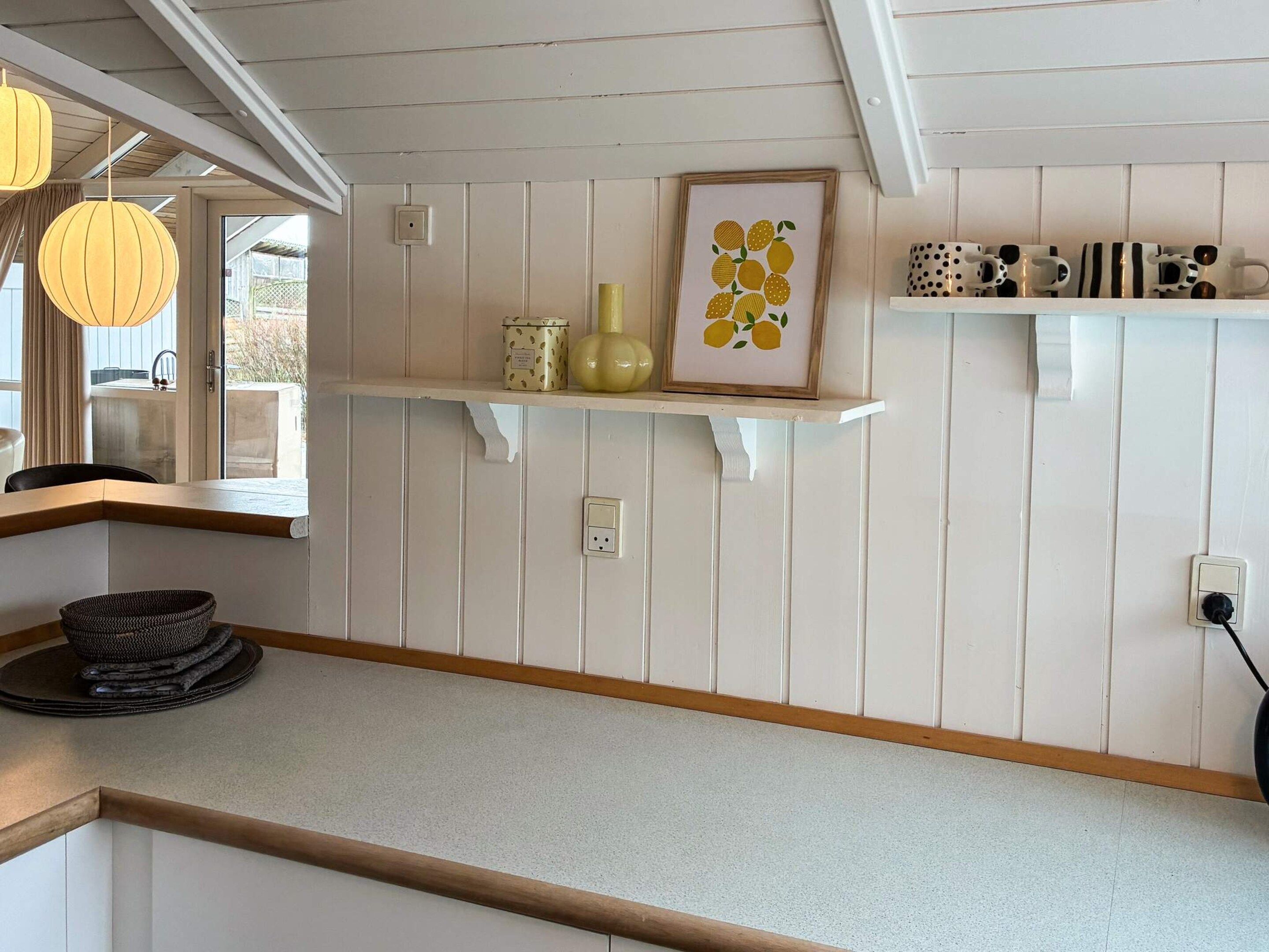 A kitchen with white wooden paneling, a countertop, a hanging light fixture, a framed picture, and a shelf with decorative items.