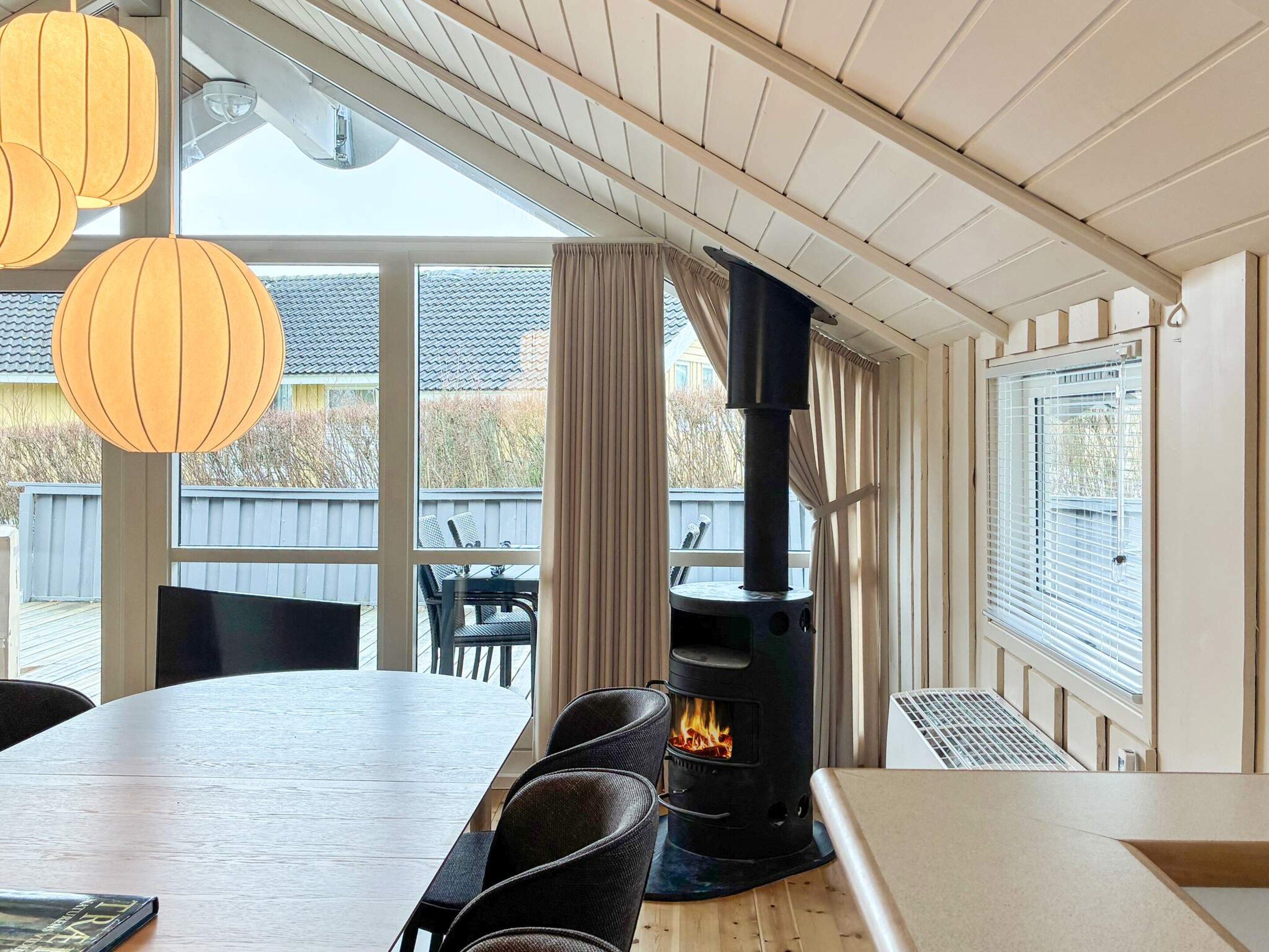 Une pièce comprenant une grande table à manger, des chaises, un poêle à bois noir et une fenêtre avec des rideaux.