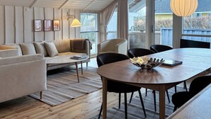 Una sala de estar moderna con sofá, mesa de comedor, sillas, ventanas amplias y lámparas colgantes.
