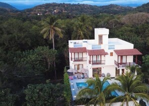 A two-story building with a swimming pool, surrounded by lush greenery and palm trees.