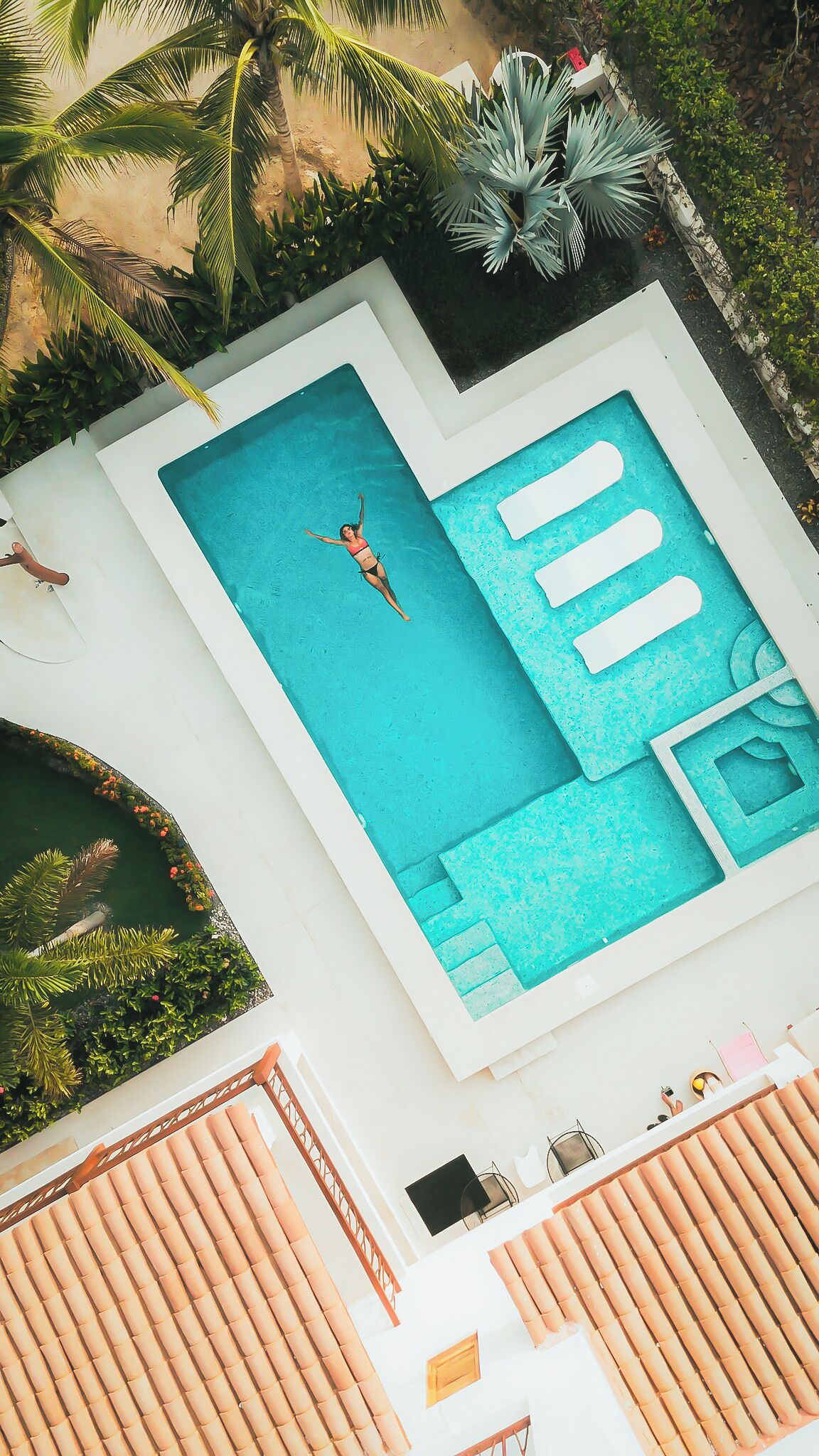 A swimming pool with a person in the water, surrounded by greenery and a white building.