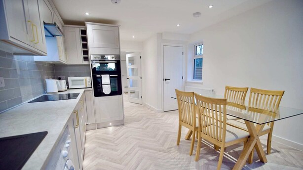 A modern kitchen with a built-in oven, microwave, and washing machine. There is a dining area with a glass table and wooden chairs. - Radcliffe N8 - 3 Bedroom House (Radcliffe)