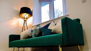 A teal sofa with gold legs, a patterned throw pillow, and a black floor lamp in a room with a window and a white wall. - Radcliffe N8 - 3 Bedroom House (Radcliffe)