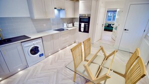 A modern kitchen with white cabinetry, a built-in oven, a coffee machine, and a washing machine. There is a glass dining table with wooden chairs. - Radcliffe N8 - 3 Bedroom House (Radcliffe)