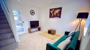 A modern living room with a herringbone floor, a green sofa, a flat-screen TV, a world map artwork, and a lamp. - Radcliffe N8 - 3 Bedroom House (Radcliffe)