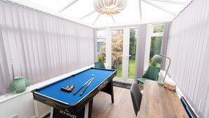 Une table de billard avec des queues et des billes, une chaise verte et une lampe dans une pièce dotée de grandes fenêtres et d’un plancher en bois.