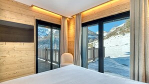 A room with wooden walls, a bed, a chair, and large windows with a mountain view.