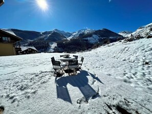 Un espace extérieur enneigé avec une table ronde et des chaises, un bâtiment en arrière-plan et des montagnes enneigées sous un ciel dégagé.