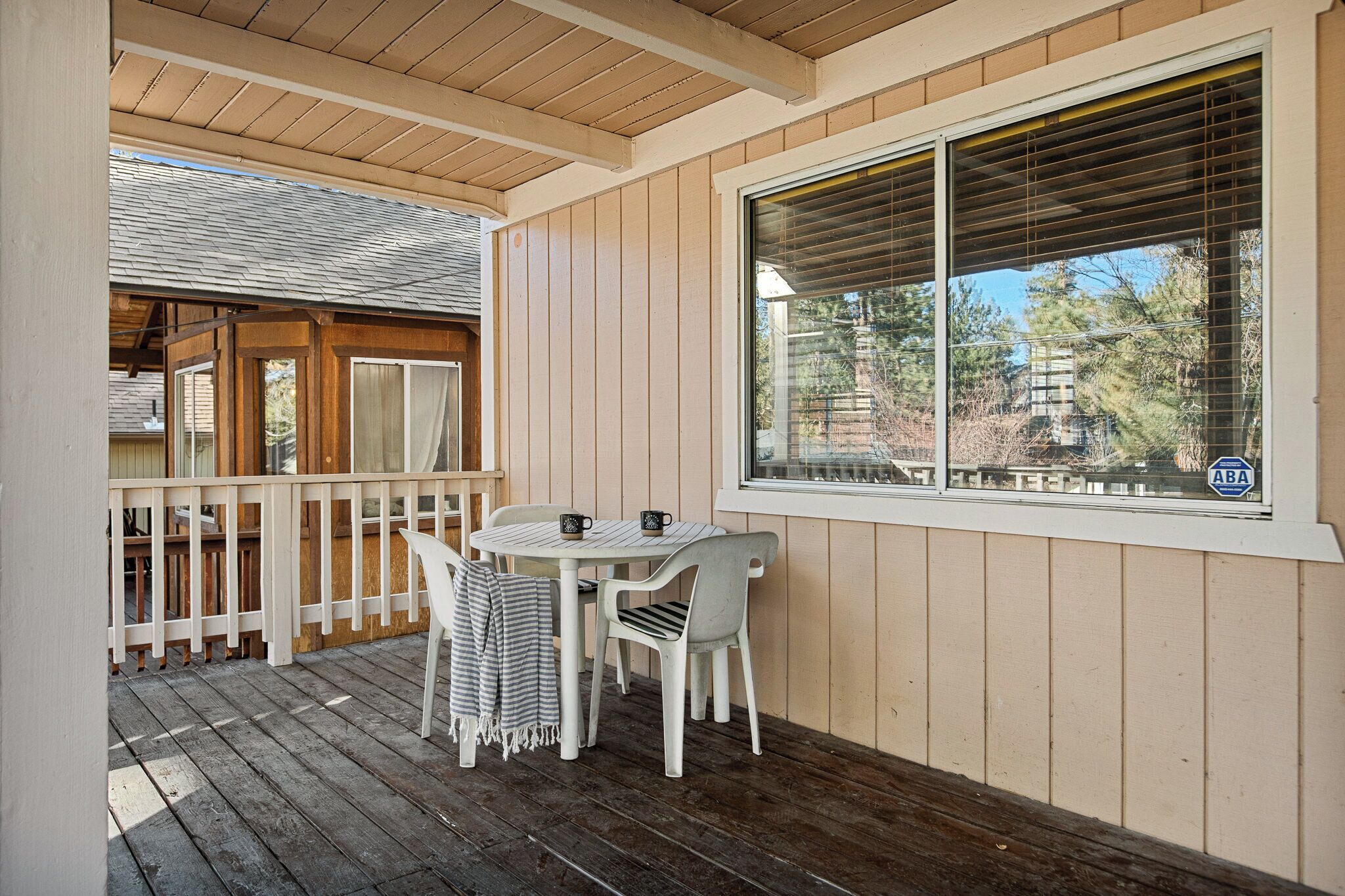 Ein überdachter Außenbereich mit Holzterrasse, einem Tisch mit Stühlen und einem Fenster mit Jalousien.