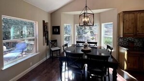 Un comedor con una mesa grande y sillas, una lámpara colgante y ventanas amplias con vistas a árboles.