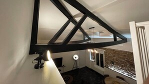 A room with a sloped ceiling, exposed beams, a desk lamp, a desk, a chair, a window with a stone wall, and a small table with a chair. - Luxury Apartment Atypical (Épinal)