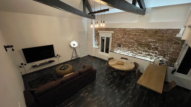 A modern living room with a dark wood floor, a round wooden table, a brown sofa, and a TV mounted on the wall. - Luxury Apartment Atypical (Épinal)