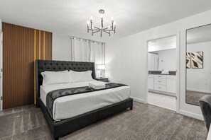 A modern bedroom with a large bed, a chandelier, a wooden panel wall, and a bathroom visible through an open door. - 5BR to Gather, Relax and Make Memories (2640ST) (Kissimmee)