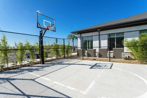 An outdoor basketball court with a chain-link fence, a basketball hoop, and a building with large windows in the background. - 5BR to Gather, Relax and Make Memories (2640ST) (Kissimmee)