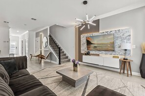 A modern living room with a sofa, a coffee table, a TV mounted on a marble wall, and a staircase. - 5BR to Gather, Relax and Make Memories (2640ST) (Kissimmee)