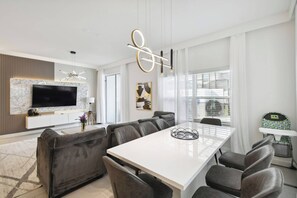 Un salon moderne doté d’un grand téléviseur à écran plat, d’une cheminée en marbre, d’un coin repas avec une table blanche et des chaises grises, ainsi que d’un espace pour changer les bébés.