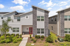 A modern two-story residential building with a stone facade, a red door, and a well-maintained front yard with shrubs and a small tree. - 5BR to Gather, Relax and Make Memories (2640ST) (Kissimmee)