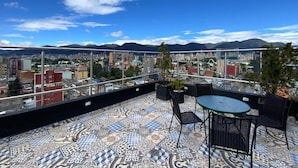 Um terraço no telhado com piso estampado, uma mesa e cadeiras, e vista para um horizonte urbano com montanhas ao fundo.