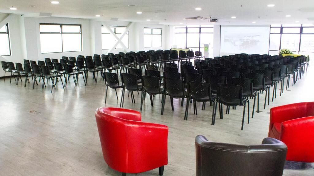 Un vaste hall avec des rangées de chaises noires et quelques chaises rouges disposées au premier plan.