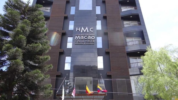 Um edifício moderno de hotel com varandas, um letreiro com a inscrição 'HMC Hotel Macao Colombia' e bandeiras hasteadas na fachada.