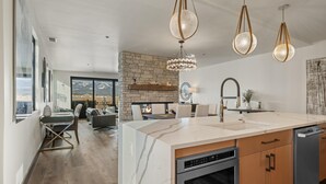 A modern kitchen with a large island, built-in oven, and stainless steel appliances. There is a stone fireplace and a dining area with a view of mountains.