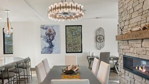 A modern dining room with a long table, white chairs, a chandelier, a fireplace, and two framed pictures on the wall.